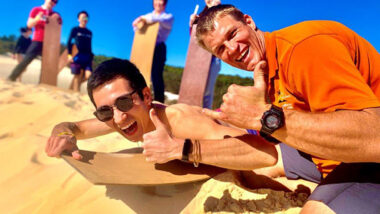Two men smiling on sand surfboard on sand hill